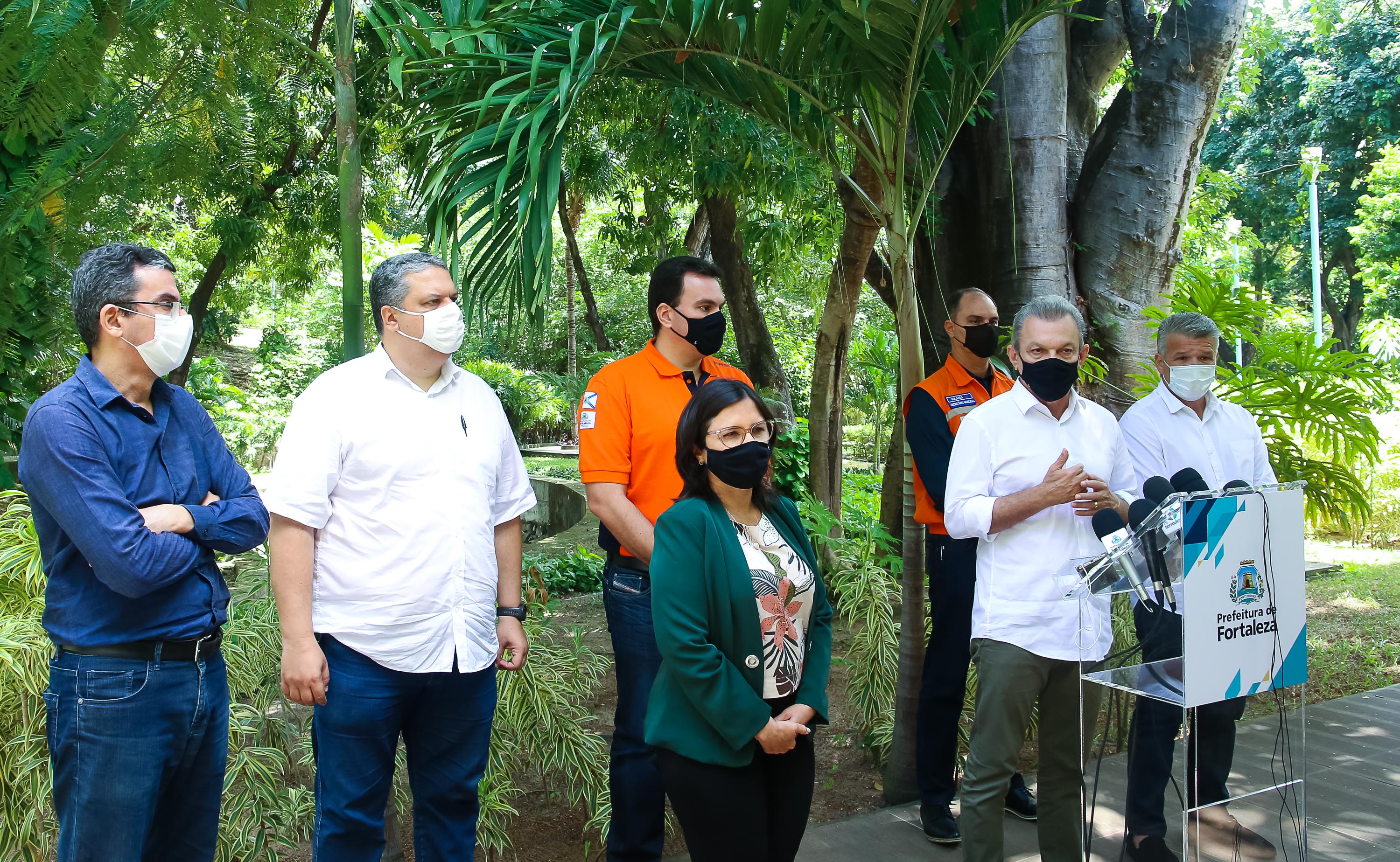 grupo de pessoas em pé em um jardim, todos usando máscara, enquanto dão entrevista